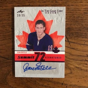 May include: A hockey card featuring Jean Ratelle, a former NHL player. The card is from the Summit 72 series and is numbered 18/35. The card has a red maple leaf design and features Ratelle's autograph.