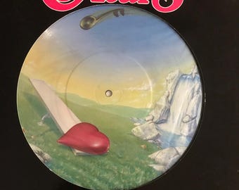 May include: A vintage vinyl record featuring the band Heart. The album cover has a black border with the band's logo in pink and white. The record's center depicts a surreal landscape with a red heart, waterfall, and a sky.