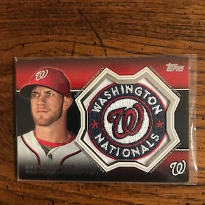 May include: A Topps baseball card featuring Bryce Harper of the Washington Nationals. The card has a commemorative patch with the team logo and text. The card is in a protective sleeve.