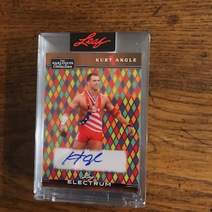 Puede incluir: Una tarjeta coleccionable de Kurt Angle de The Harlequin Collection, en un estuche protector transparente. La tarjeta presenta a Kurt Angle con atuendo de lucha libre, con un tema de la bandera estadounidense. El fondo tiene un patrón de diamantes colorido y está firmado.