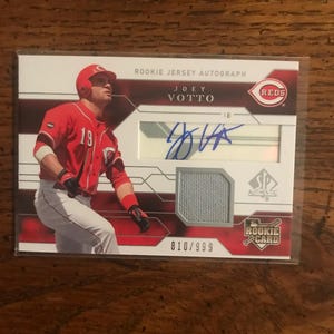 Puede incluir: Una tarjeta de béisbol Joey Votto Rookie Jersey Autograph. La tarjeta presenta una foto de Votto con el uniforme rojo de los Rojos de Cincinnati. La tarjeta incluye una muestra de camiseta y su firma. La tarjeta está numerada 810/999.