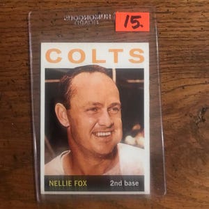 Puede incluir: Una tarjeta coleccionable deportiva vintage con un hombre sonriente y la palabra "COLTS" en naranja. La tarjeta está etiquetada como "NELLIE FOX 2nd base" y está en una funda protectora. Una etiqueta de precio roja con el número "15" es visible.
