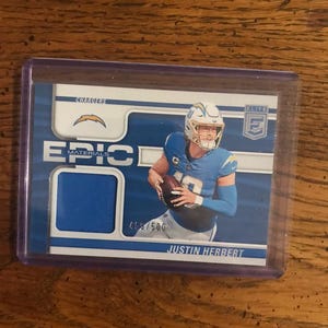 May include: A blue and white football trading card featuring Justin Herbert of the Los Angeles Chargers. The card has a blue jersey swatch and the text "EPIC MATERIALS". The card is encased in a clear plastic holder.