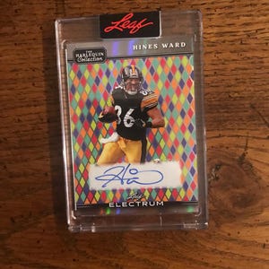 May include: A Hines Ward football trading card from The Harlequin Collection, encased in a clear protective holder. The card features a photo of Ward in his Steelers uniform, with a colorful diamond pattern background and his signature.