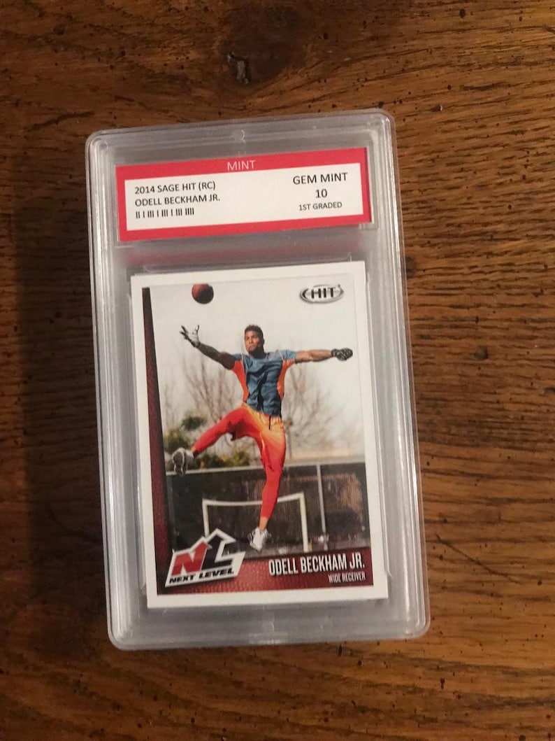 May include: A graded sports card featuring Odell Beckham Jr. in a red and orange uniform, holding a football. The card is graded GEM MINT 10 and is from the 2014 Sage Hit series.