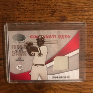 Puede incluir: Una tarjeta de béisbol con un jugador con un uniforme blanco con rayas rojas y marrones, sosteniendo un bate. La tarjeta tiene un fondo rojo con el texto "Cincinnati Reds" y "Fabric of the Game" en blanco. El nombre del jugador es "Joe Morgan".