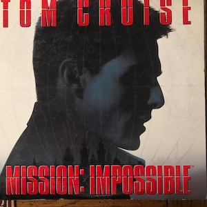 May include: A DVD case featuring Tom Cruise with the text "Tom Cruise" in red block letters at the top. The title "Mission: Impossible" is displayed in red block letters at the bottom, with a silhouette of a man's face in the center.
