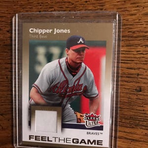 Puede incluir: Una tarjeta de béisbol con Chipper Jones de los Bravos de Atlanta. La tarjeta tiene un parche de tela y el texto "FEEL THE GAME". La tarjeta está en una funda protectora.