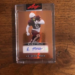 May include: A football trading card encased in a clear protective holder. The card features a player in a maroon and white uniform, with the name "LE'VEON MOSS" and a signature. The card is labeled "ELECTRUM" and "LEAF".