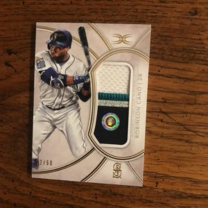 Puede incluir: Tarjeta de béisbol con un jugador con uniforme de béisbol, sosteniendo un bate. La tarjeta incluye un parche de tela y el nombre del jugador, Robinson Cano, y el número 2/50.