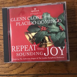 May include: A CD case with a red cover featuring the text "GLENN CLOSE and PLACIDO DOMINGO" and "REPEAT THE JOY". The cover art includes gold bells, holly leaves, and red berries. The Hallmark logo is in the upper left corner.
