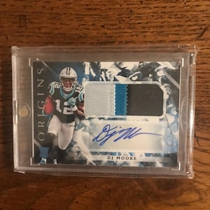 May include: A sports trading card featuring a player in a Carolina Panthers uniform. The card has a patch of fabric with black, white, and teal colors. The card is signed "DJ Moore" and is encased in a clear plastic holder.