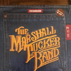 Puede incluir: Una portada de álbum de vinilo con un fondo de mezclilla y un logotipo bordado en amarillo que dice "The Marshall Tucker Band". La portada del álbum tiene una pegatina que dice "Tuckerized" y otra pegatina que dice "Record Bar 8.49".