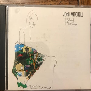 Puede incluir: Una caja de CD blanca con una línea negra en el lado izquierdo. La parte frontal de la caja presenta un dibujo en blanco y negro de una mujer con una ilustración colorida de una casa y un coche detrás de ella. El texto "JONI MITCHELL" está impreso en negro en la esquina superior derecha de la caja. El texto "Ladies of The Canyon" está impreso en negro debajo del nombre del artista.