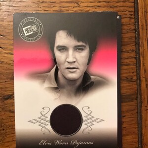 May include: A black and white photo of Elvis Presley on a card with a red and pink background. The card has a black circle with a piece of fabric inside. The text on the card reads "Press Pass Authentic", "Elvis Worn Pajamas".