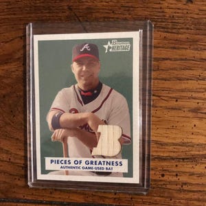 May include: A baseball card featuring a player in a white and red uniform with the text "PIECES OF GREATNESS." The card is in a protective sleeve. The player is wearing a baseball cap with the letter "A."