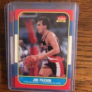 Puede incluir: Una tarjeta de baloncesto de Jim Paxson de la serie Fleer Premier. La tarjeta presenta una foto de Paxson con el uniforme de los Trail Blazers, sosteniendo un balón de baloncesto. La tarjeta está dentro de una funda protectora.