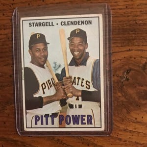 Puede incluir: Una tarjeta de béisbol vintage con Stargell y Clendenon de los Piratas de Pittsburgh, sosteniendo bates. La tarjeta tiene un borde blanco con el texto "PITT POWER" y el nombre del equipo. Los jugadores llevan gorras negras con un logotipo "P".