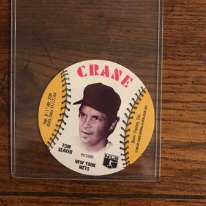 May include: A white baseball card with a yellow border featuring a baseball player wearing a white and black baseball uniform. The card has the text "CRANE" at the top, "TOM SEAVER" in the bottom left corner, "PITCHER" in the bottom center, "NEW YORK METS" in the bottom right corner, and "MLB" in the bottom center. The card also has the text "Hgt. 6'-1" Wt. 206 Birth Date 11/17/44" in the top left corner and "Born: Fresno, Cal." in the top right corner.