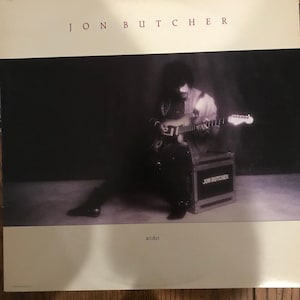 May include: A black and white photo of a musician playing a guitar. The musician is sitting on a box with the words "Jon Butcher" printed on it. The album cover is titled "Wishes" and features the artist's name "Jon Butcher" at the top.