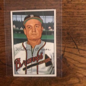 May include: A vintage baseball card featuring Walker Cooper of the Braves. The card shows a portrait of Cooper in his team uniform, with the team name in red script. The background depicts a baseball field.