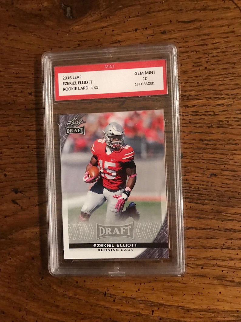 K&ouml;nnte beinhalten: Eine bewertete Sportkarte mit Ezekiel Elliott, einem Footballspieler, in einem rot-wei&szlig;en Trikot. Die Karte stammt aus der Leaf Draft-Serie von 2016 und ist mit Gem Mint 10 bewertet.