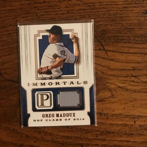 Könnte beinhalten: Eine Baseballkarte mit Greg Maddux in einer Wurfpose. Die Karte hat ein blaues und weißes Design mit dem Text "IMMORTALS" oben und "GREG MADDUX HOF CLASS OF 2014" unten. Die Karte enthält auch einen Patch.