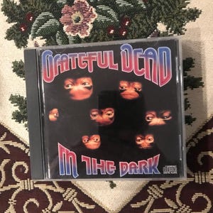 May include: A compact disc case featuring the album "In the Dark" by The Grateful Dead. The album cover has the band's name in a red and blue font, with multiple eyes on a black background. The disc is a digital audio disc.
