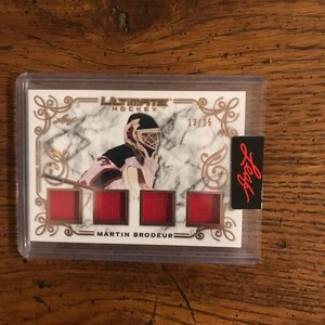 May include: A white hockey card with a gold border and a picture of a hockey player wearing a red and white jersey. The card has the text "Ultimate Hockey" and "Martin Brodeur" on it. The card is numbered 13/25.