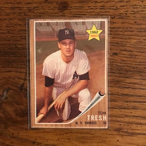Puede incluir: Una tarjeta de béisbol vintage con Tom Tresh, un jugador de los New York Yankees. La tarjeta es de 1962 y presenta una foto de Tresh con su uniforme, sosteniendo un bate. La tarjeta tiene una estrella amarilla con el texto "1962 Rookie" en ella.