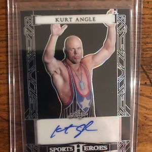 Puede incluir: Una tarjeta de deporte negra y plateada con una foto de Kurt Angle, un luchador profesional, con un maillot de lucha. La tarjeta tiene un panel de firma blanco con una firma azul y el texto "SPORTS HEROES".