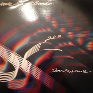 Puede incluir: Una portada de álbum de vinilo con fondo negro y un diseño abstracto colorido. El álbum se titula "Time Exposure" de "Little River Band".