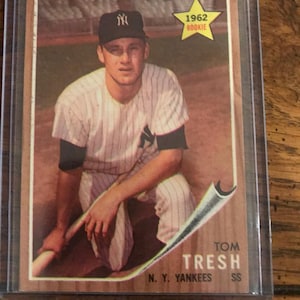 Puede incluir: Una tarjeta de béisbol vintage con Tom Tresh de los Yankees de Nueva York. La tarjeta muestra a Tresh con su uniforme de los Yankees, con una gorra negra y un emblema de "1962 Rookie" en forma de estrella. La tarjeta está en una funda protectora.