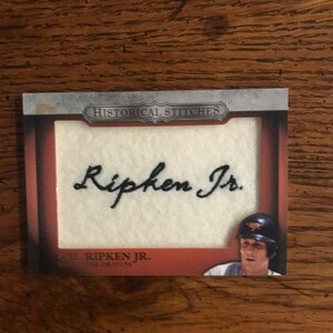 Pode incluir: Um cartão de beisebol com a assinatura bordada de Ripken Jr. O cartão tem um fundo creme e as palavras "Historical Stitches". Uma pequena imagem de um jogador de beisebol com capacete é visível.
