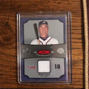 Könnte beinhalten: Baseballkarte mit Chipper Jones von den Atlanta Braves. Die Karte zeigt Jones in seiner Braves-Uniform mit einem Schläger. Sie enthält ein spielgebrauchtes Stoffmuster und die Nummer 10. Die Karte ist in einem Schutzhalter eingeschlossen.