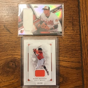 May include: Two baseball cards featuring Manny Machado of the Baltimore Orioles. The top card is a silver and red "Out Gear" card with a jersey patch. The bottom card is a red, white, and gold "National Treasures" card with a jersey patch and the number 43/99.