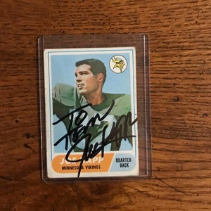 May include: A vintage football trading card featuring Joe Kapp of the Minnesota Vikings. The card has a blue and white background with a portrait of Kapp in his green uniform. The card is signed in black ink and encased in a protective sleeve.
