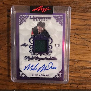 May include: A purple and white hockey card featuring Mike Modano, a former NHL player. The card is a Leaf Ultimate Hockey card with an autographed jersey patch and the number 6/10. The card is in a plastic case.