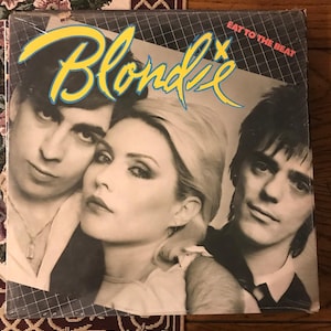 May include: Blondie's "Eat to the Beat" album cover. The cover features three band members in a black and white photo. The band's name is in yellow script with a blue outline. The album title is in red text.