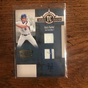 Puede incluir: Tarjeta de béisbol de Gary Carter de los Mets de Nueva York. La tarjeta tiene un diseño azul y blanco con las palabras "Throwback Collection" y "TC". La tarjeta está dentro de una funda de plástico transparente.