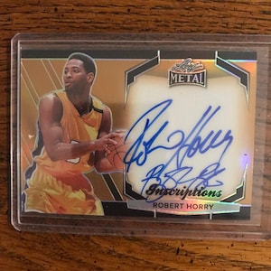 Op de afbeelding: Een basketbal trading card met Robert Horry in een goudgeel uniform, met een basketbal. De kaart heeft een gouden rand en de tekst "LEAF METAL" bovenaan. De kaart is met blauwe inkt gesigneerd.