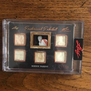 Può includere: Una carta da hockey in bianco e nero con una foto di Bernie Parent in uniforme da hockey. La carta fa parte della serie Enshrined Exhibit Art of Hockey ed è numerata 17/35.