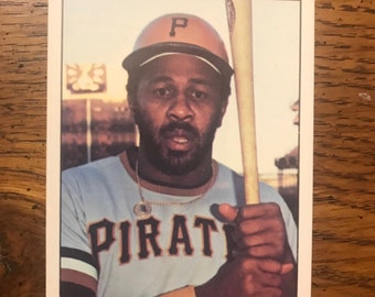 Könnte beinhalten: Eine Baseballkarte mit einem Baseballspieler, der eine blaue und braune Uniform mit dem Wort "PIRATE" auf der Vorderseite trägt. Der Spieler hält einen Holzschläger und trägt eine braune Baseballkappe mit einem weißen "P" auf der Vorderseite.