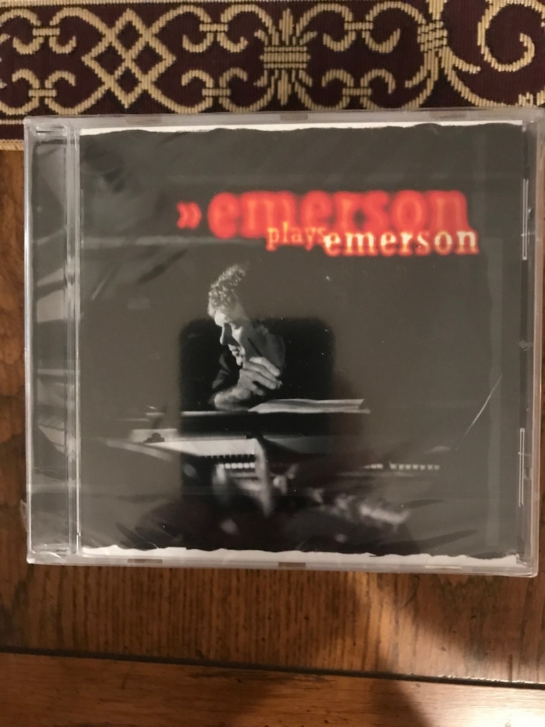 May include: A CD case featuring a black and white photograph of a man playing a keyboard. The title of the CD is "Emerson plays Emerson" in red text.