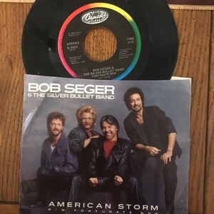 May include: A 45 RPM vinyl record single by Bob Seger & The Silver Bullet Band. The record is black with a rainbow colored label. The label features the Capitol Records logo and the song titles "American Storm" and "Fortunate Son".