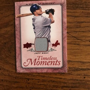 Può includere: Una carta di baseball con un giocatore di baseball in uniforme blu e bianca che swinga una mazza. La carta ha un design rosso, bianco e blu con il testo "A Piece of History" e "Timeless Moments". La carta presenta un pezzo della maglia del giocatore.