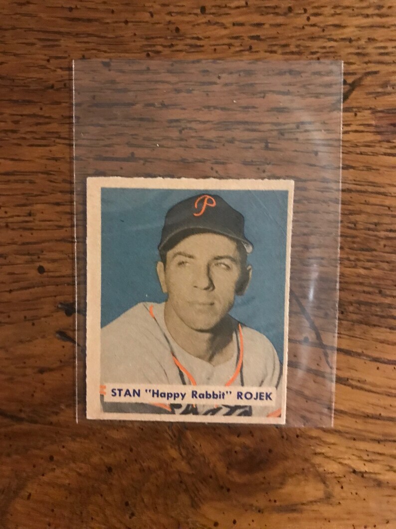 May include: A vintage baseball card featuring a color photo of Stan "Happy Rabbit" Rojek, a baseball player wearing a Philadelphia Phillies uniform. The card has a blue background and the text "STAN "Happy Rabbit" ROJEK" is printed in white at the bottom.