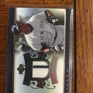 Può includere: Una carta di baseball con un giocatore di baseball che indossa una divisa bianca e grigia con la scritta "NEW YORK" sul davanti. La carta ha un bordo argentato e nero con una toppa argentata e bianca al centro. La scritta "TIGERS" è stampata sul lato destro della carta.