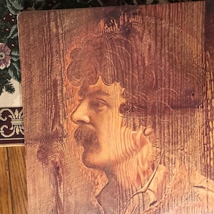 May include: A wood-grain print of a man's face with a mustache and curly hair. The print is on a wooden background.