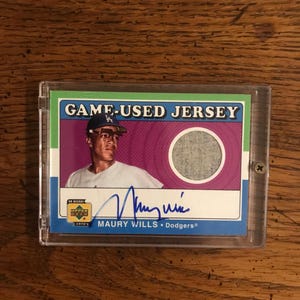 Könnte beinhalten: Eine blaue, grüne und weiße Baseballkarte mit einem Foto eines Baseballspielers, der ein weißes Trikot und eine blaue Mütze mit einem weißen LA-Logo trägt. Die Karte trägt den Text "GAME-USED JERSEY" und "MAURY WILLS - Dodgers" sowie ein blaues und gelbes Upper Deck-Logo. Die Karte enthält auch ein kleines graues Stoffstück.
